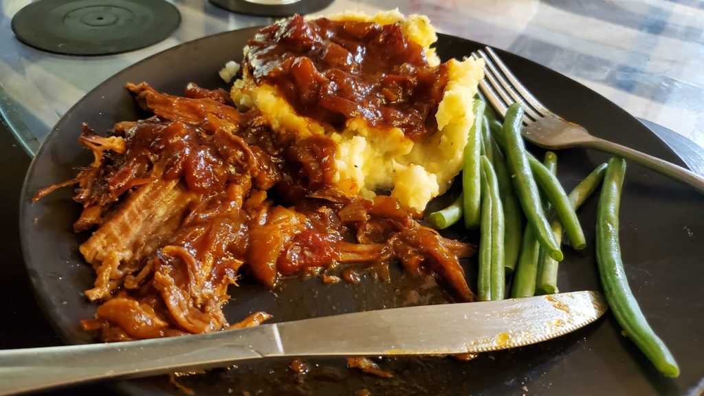 A dinner plate with beef brisket, mashed potatoes topped with the barbecue braising sauce, and string beans